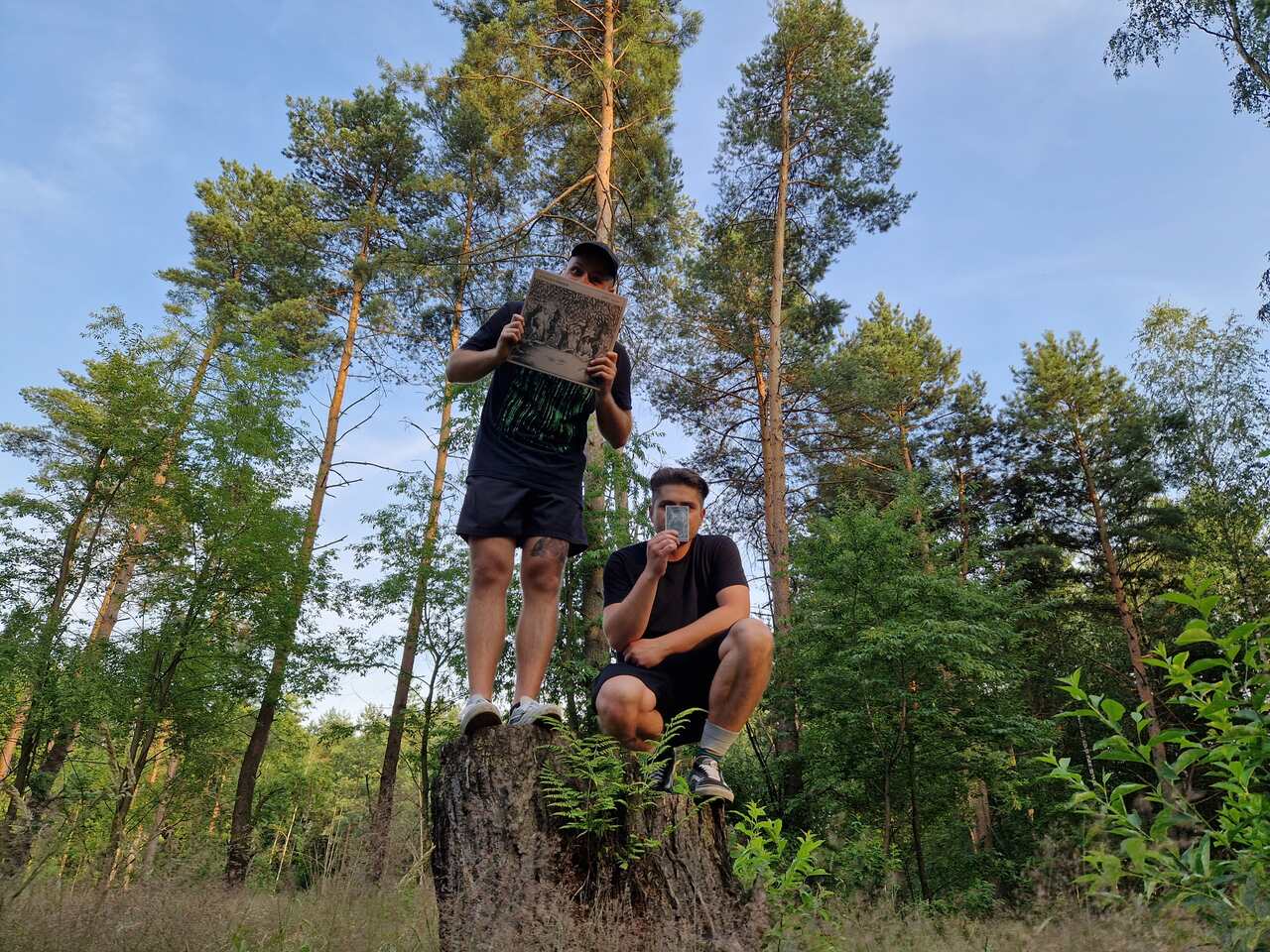 Na zdjęciu widać dwóch mężczyzn w lesie. Jeden kuca, a drugi stoi na ściętym pniu drzewa. 