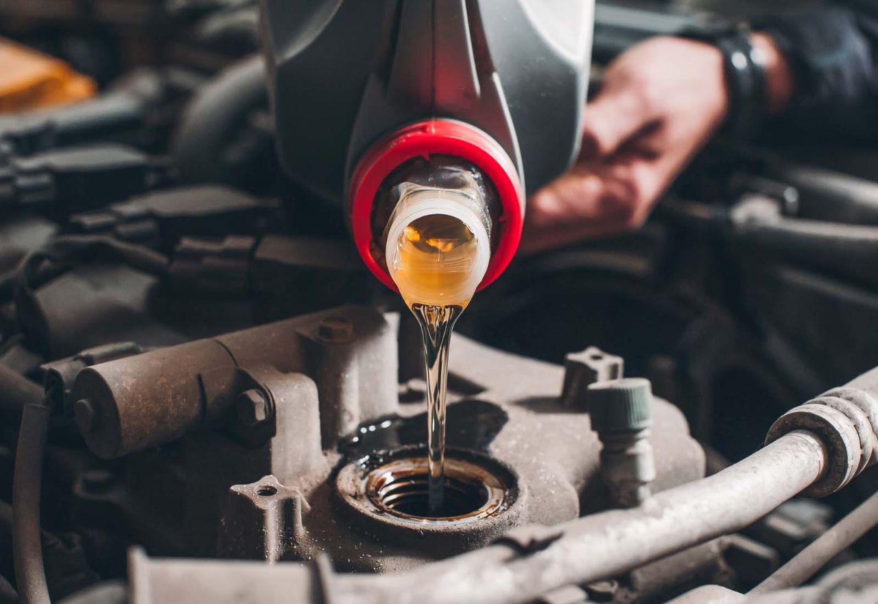 Pouring clean engine oil into a diesel truck engine during maintenance, with visible hoses, components, and technician's hand in background.