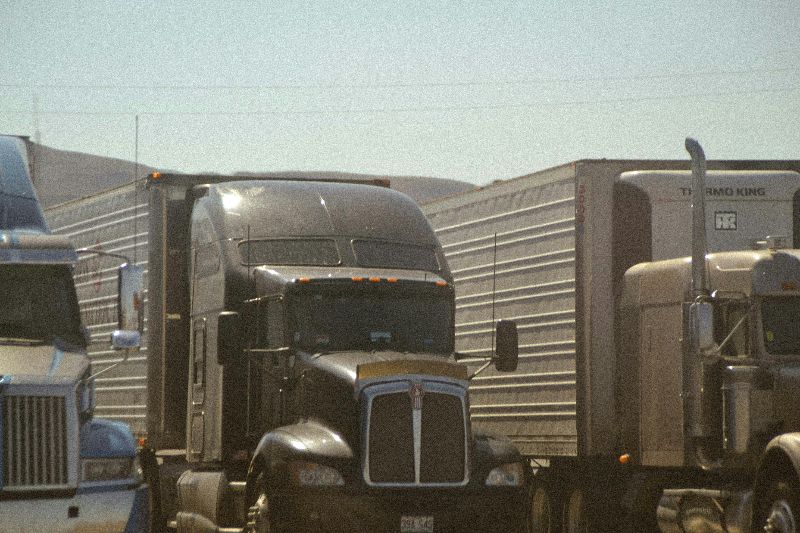 Three semi trucks drive closely together on busy highway, hauling box trailers through desert landscape under hazy afternoon sunlight conditions.