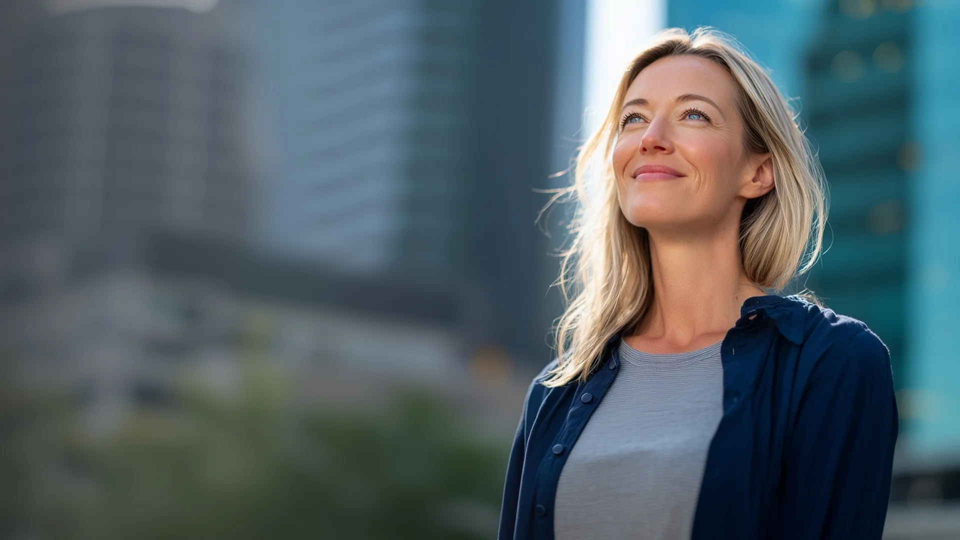 Smiling blonde woman looking upward with city buildings blurred in the background.