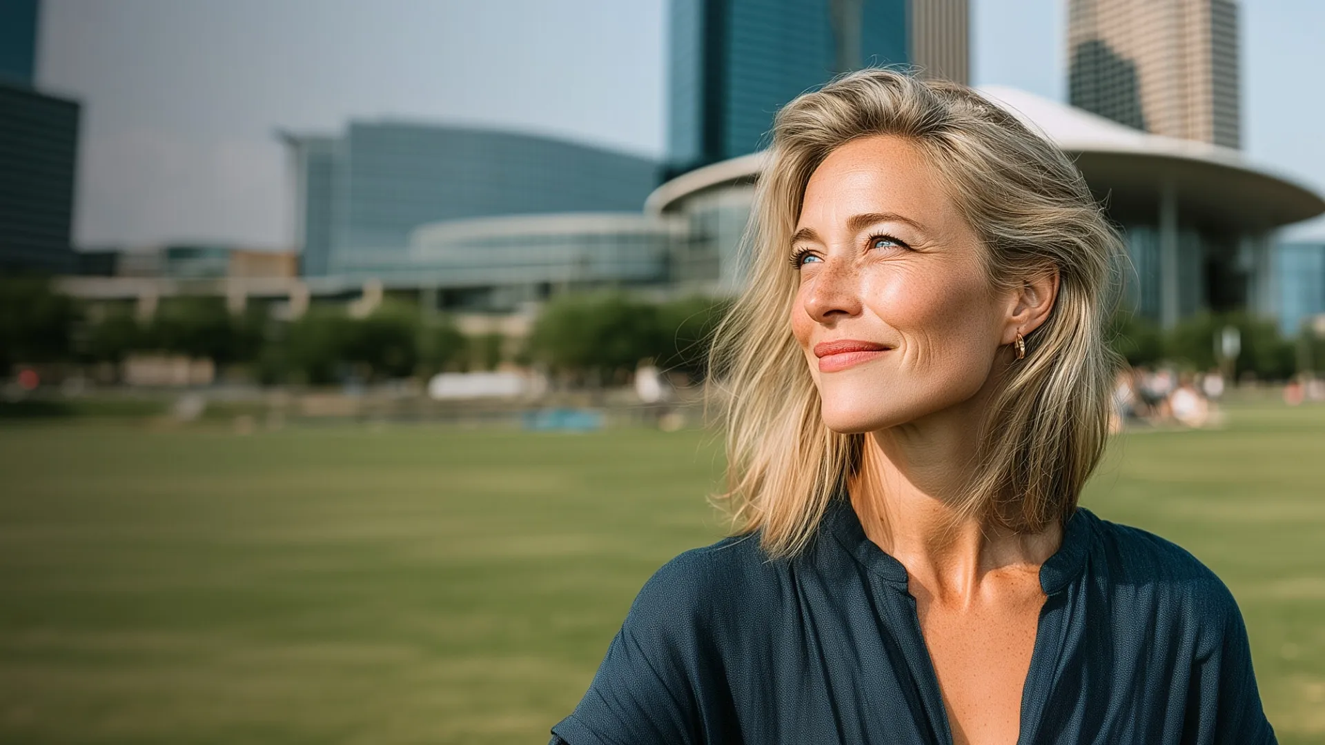 Blonde woman smiling outdoors with modern buildings and green lawn in the background.