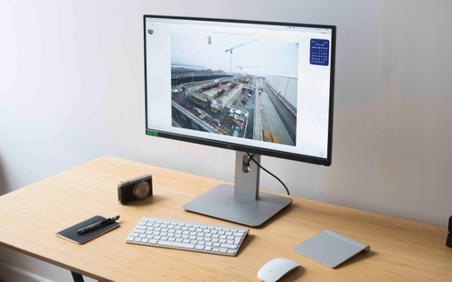 Office desk with keyboard, mouse, camera, notebook with pen, and monitor showing a construction site image.