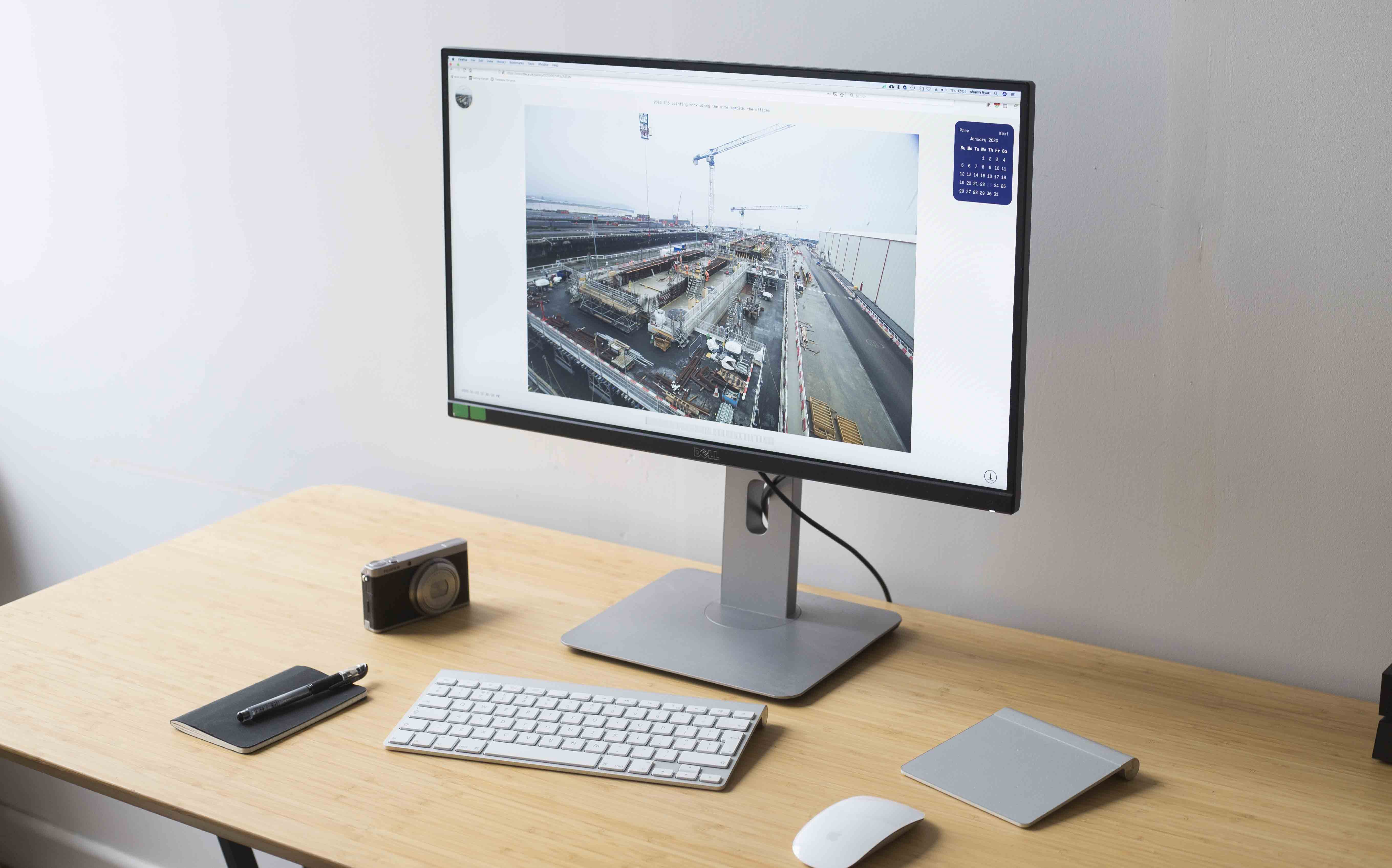 Office desk with keyboard, mouse, camera, notebook with pen, and monitor showing a construction site image.