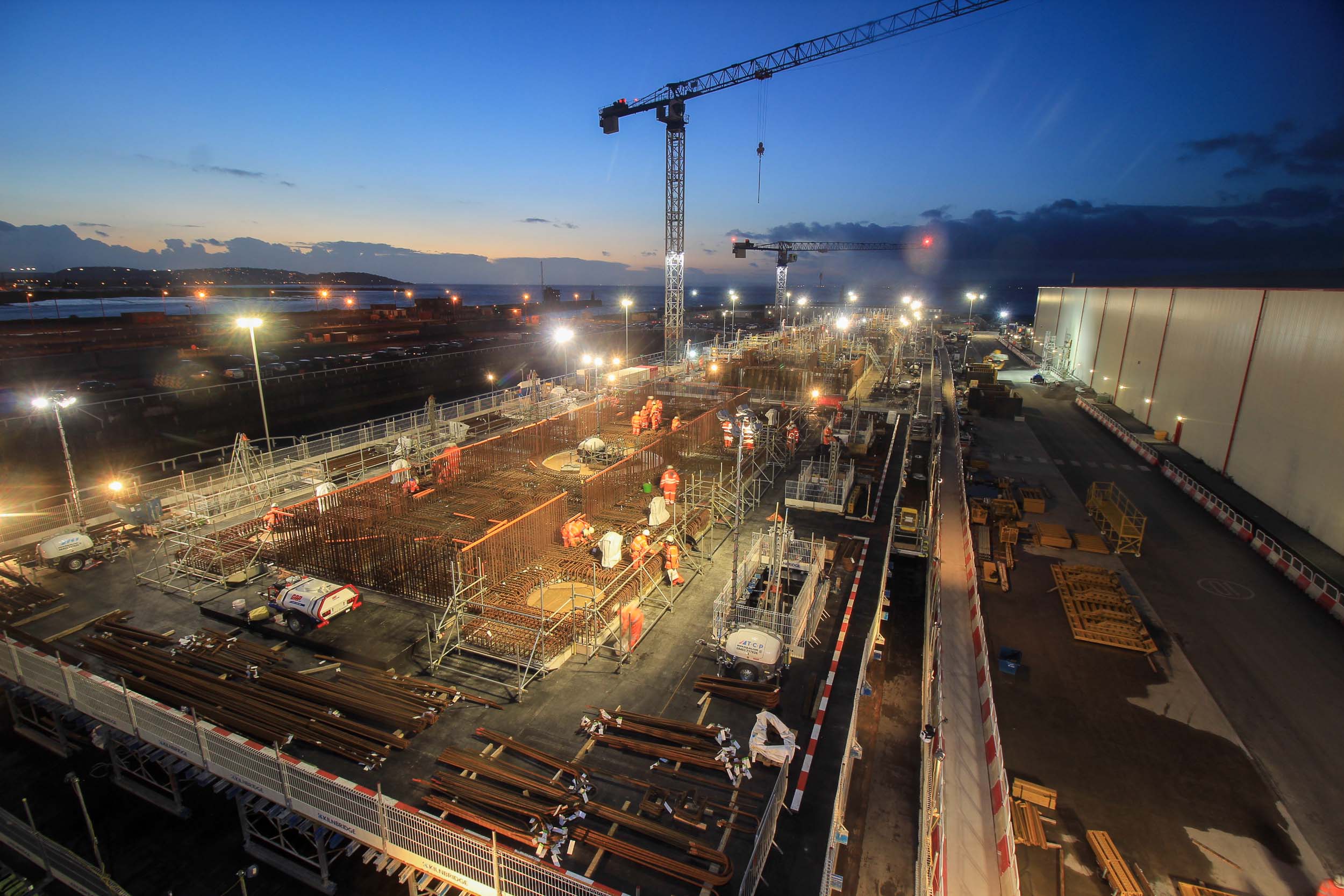 Night view of a large construction site with cranes, workers, and building materials under bright lights.
