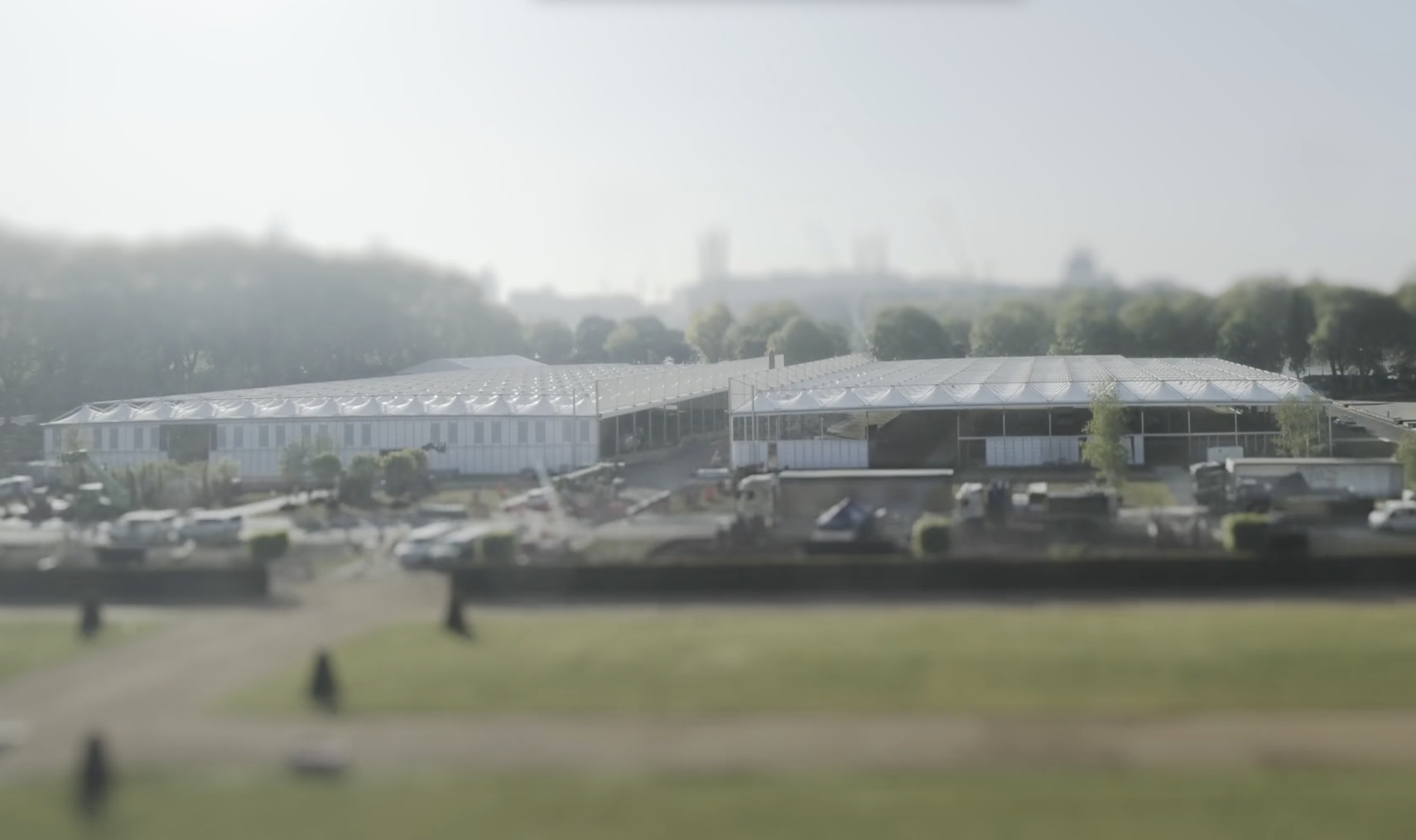 Two large white industrial buildings with solar panels on the roofs seen from a distance with trees behind.