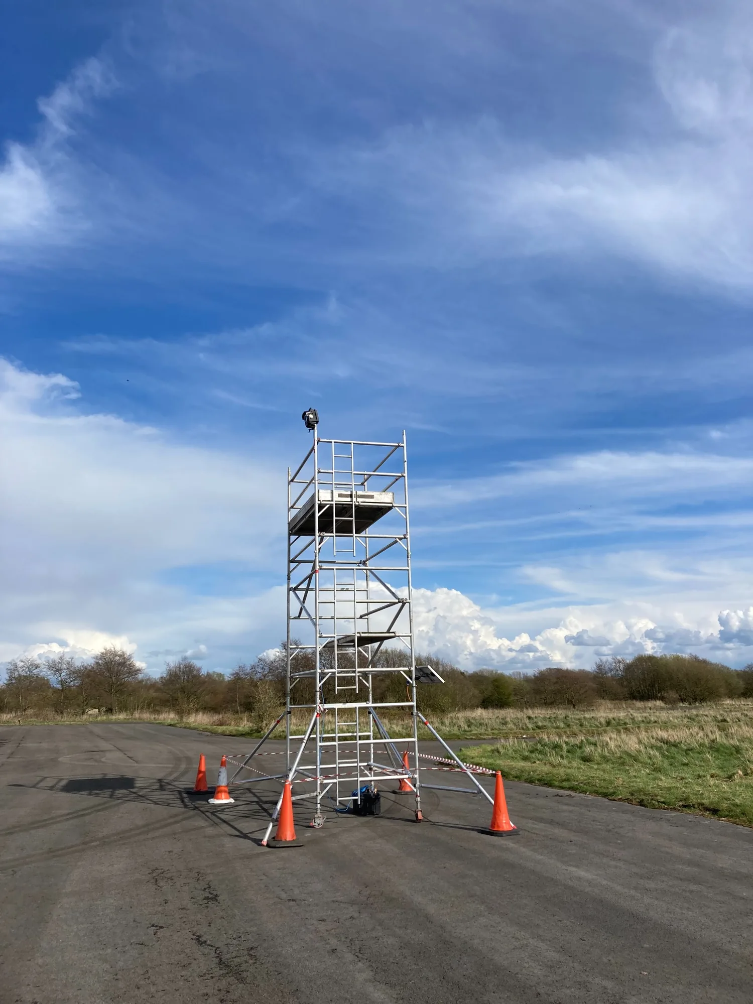 Metal scaffold tower with safety cones on an asphalt area with grass and trees under blue sky.