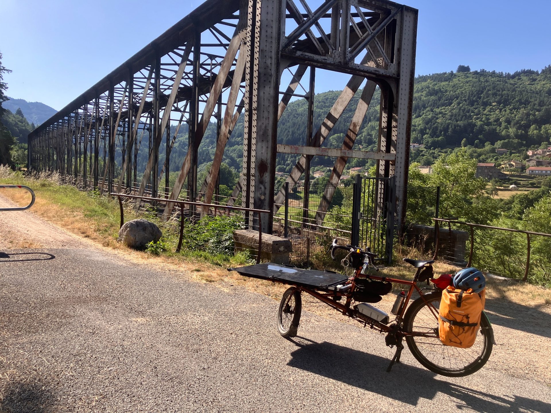 Cargo bike with orange pannier and blue helmet parked near rusty metal bridge in rural area.