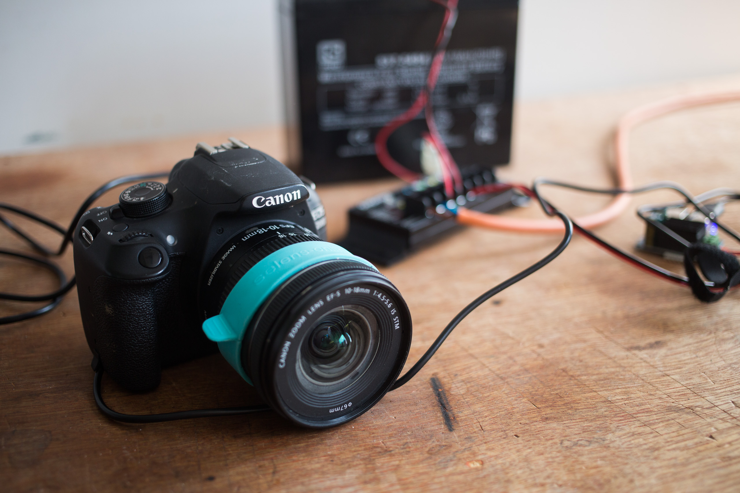 Black Canon DSLR camera with a turquoise lens attachment on a wooden surface, connected to wires and electronics.