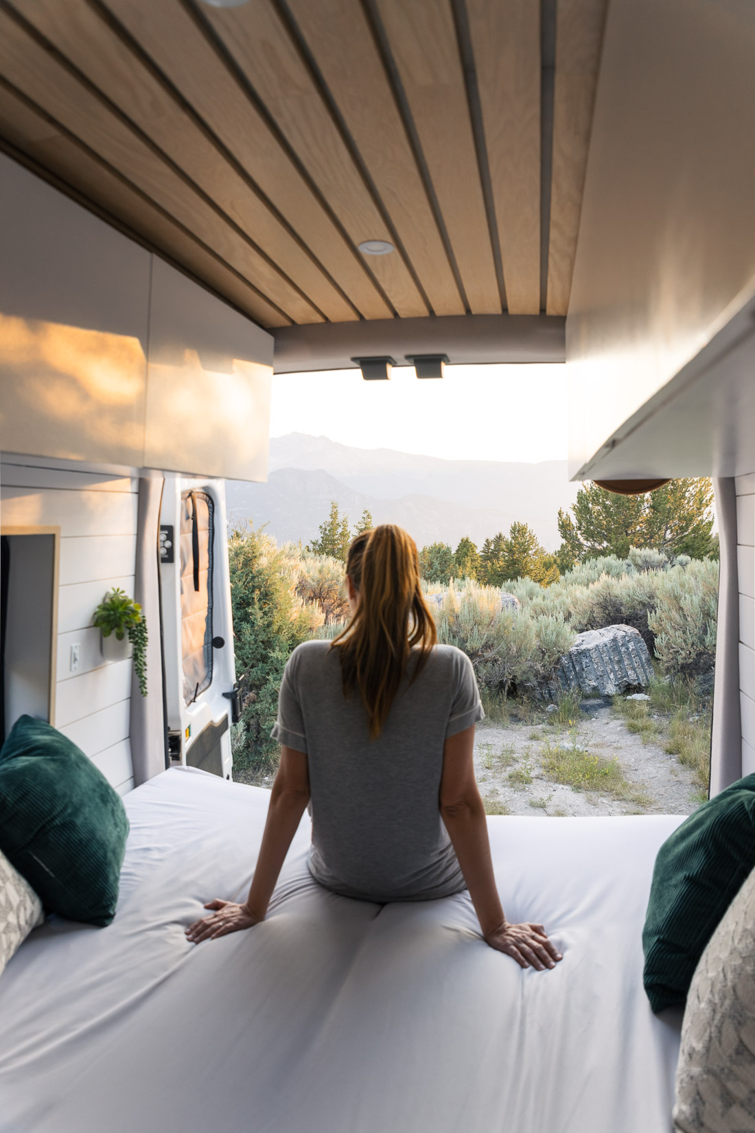 A woman sits on the edge of a bed in the back of a campervan with the doors open to a view of plants and a mountain view during sunset.