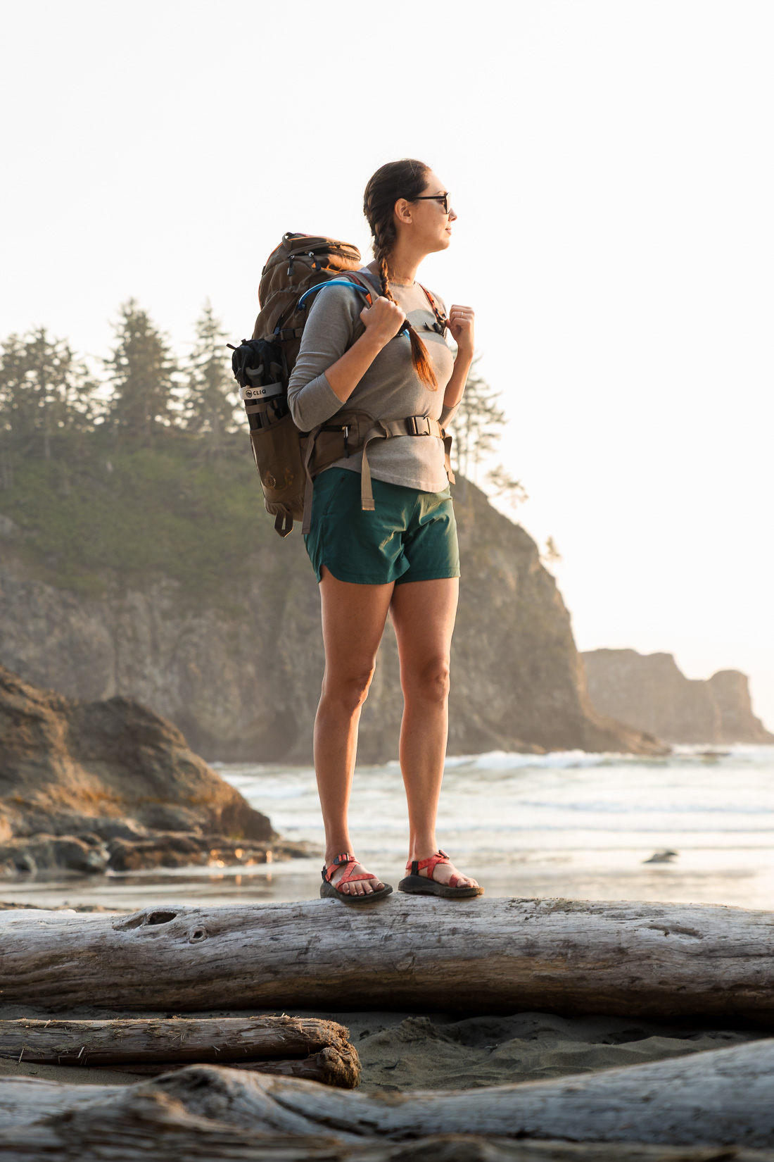 A woman wearing a backpack stands on a log in front of a Pacific Northwest beach during a golden sunset.
