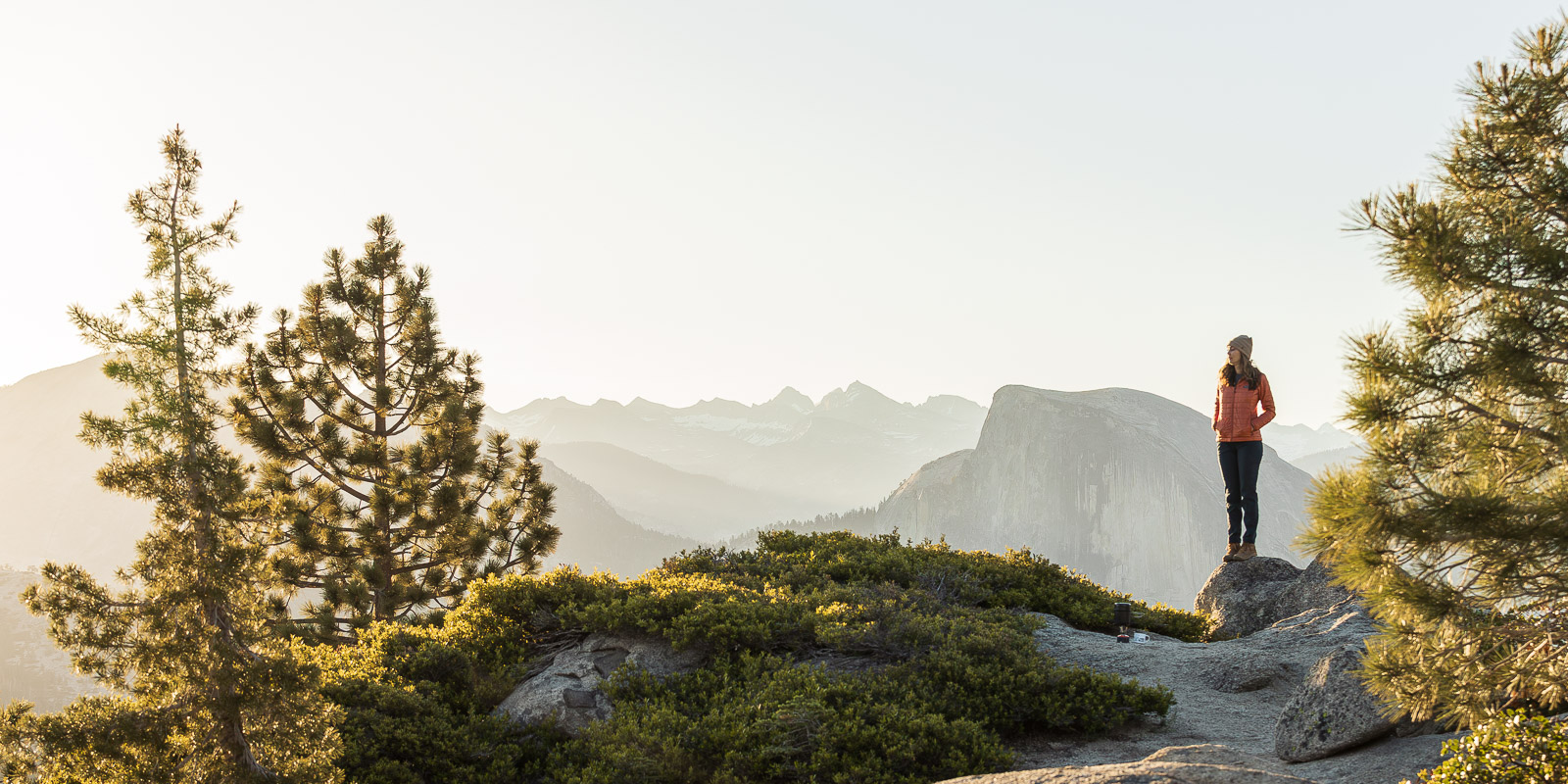 A woman stands on a rock during sunrise with pine trees on either side and an expansive view of Half Dome and other mountain peaks in the distance.