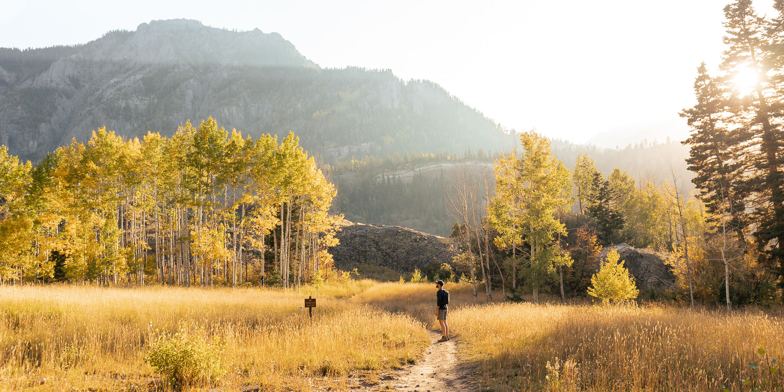 A man standing on a dirt trail that passes through golden aspen trees with mountains in the background during a sunset