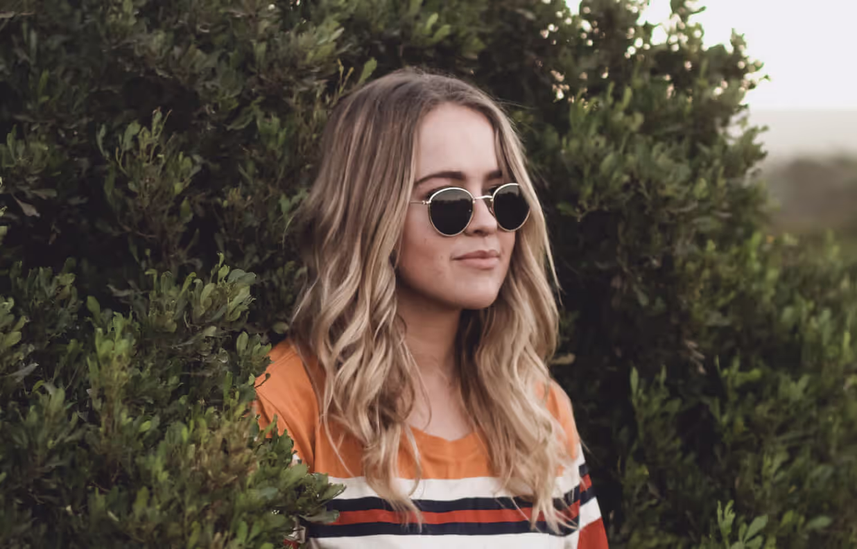 A woman with blonde hair and sunglasses standing in front of a bush.