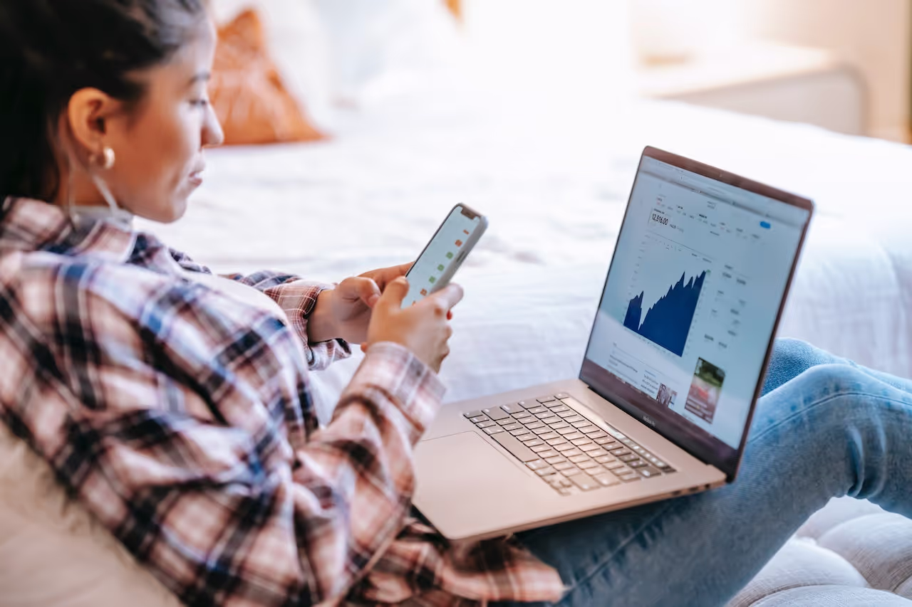 A woman holding a cell phone and a laptop analyzing stocks charts