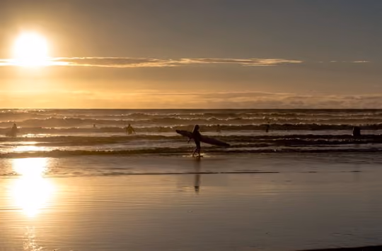 Surfer walking with board in hand as the sun sets over the ocean behind them