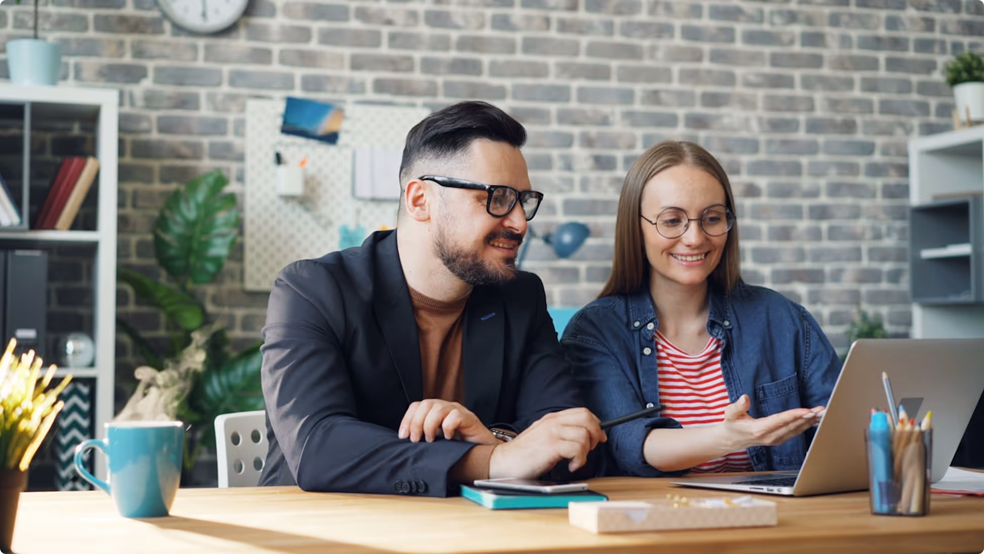 Two people wearing glasses smiling and looking at a laptop screen in a modern office with brick walls.