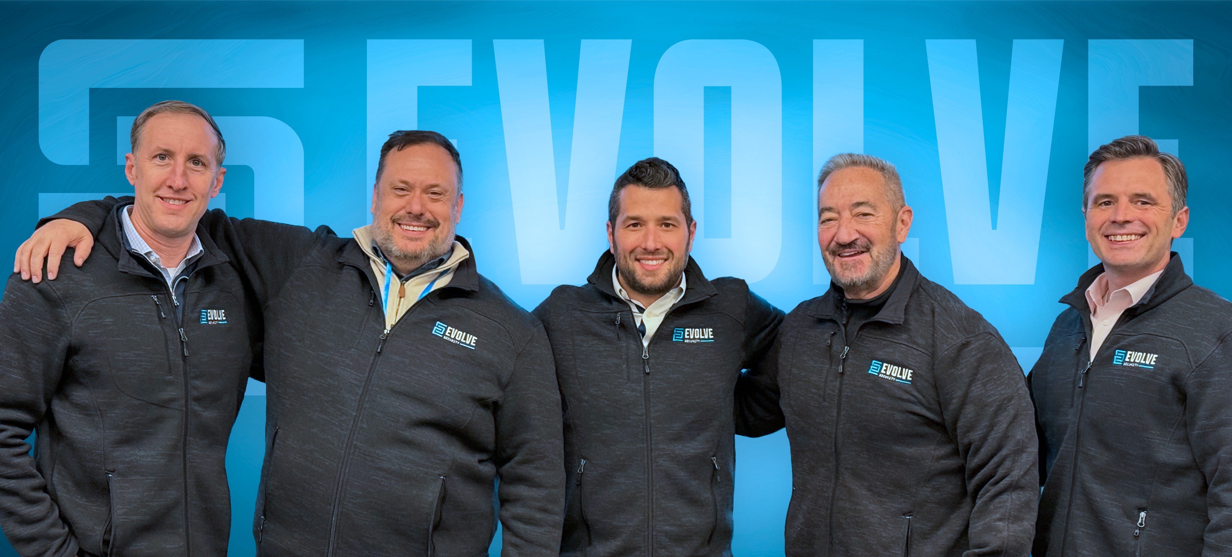 Four men wearing matching blue and black North Face jackets with Evolve Security logos, standing indoors in front of plants and a screen.