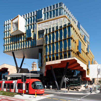 Modern multi-story building with glass and gold panels by a street with a red tram and pedestrians.