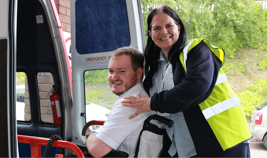 Smiling woman in safety vest supports man seated in wheelchair by an open vehicle door.