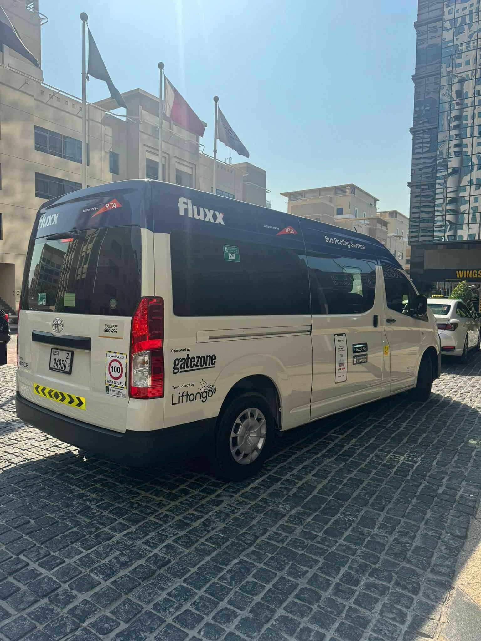 White and blue Fluxx bus pooling van parked on cobblestone street in urban area under sunlight.