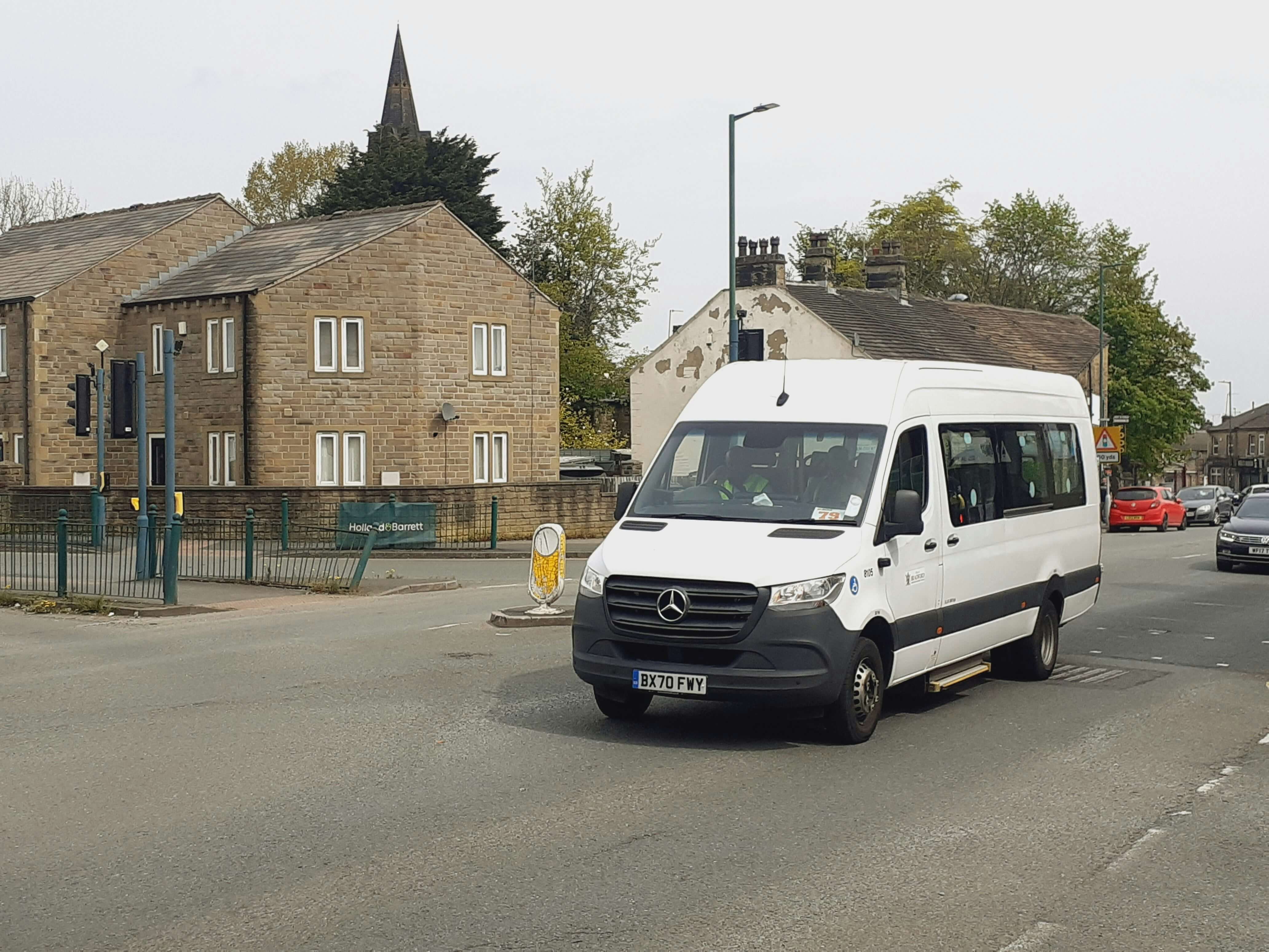 White Mercedes van driving on a street in a residential area with stone houses and trees.