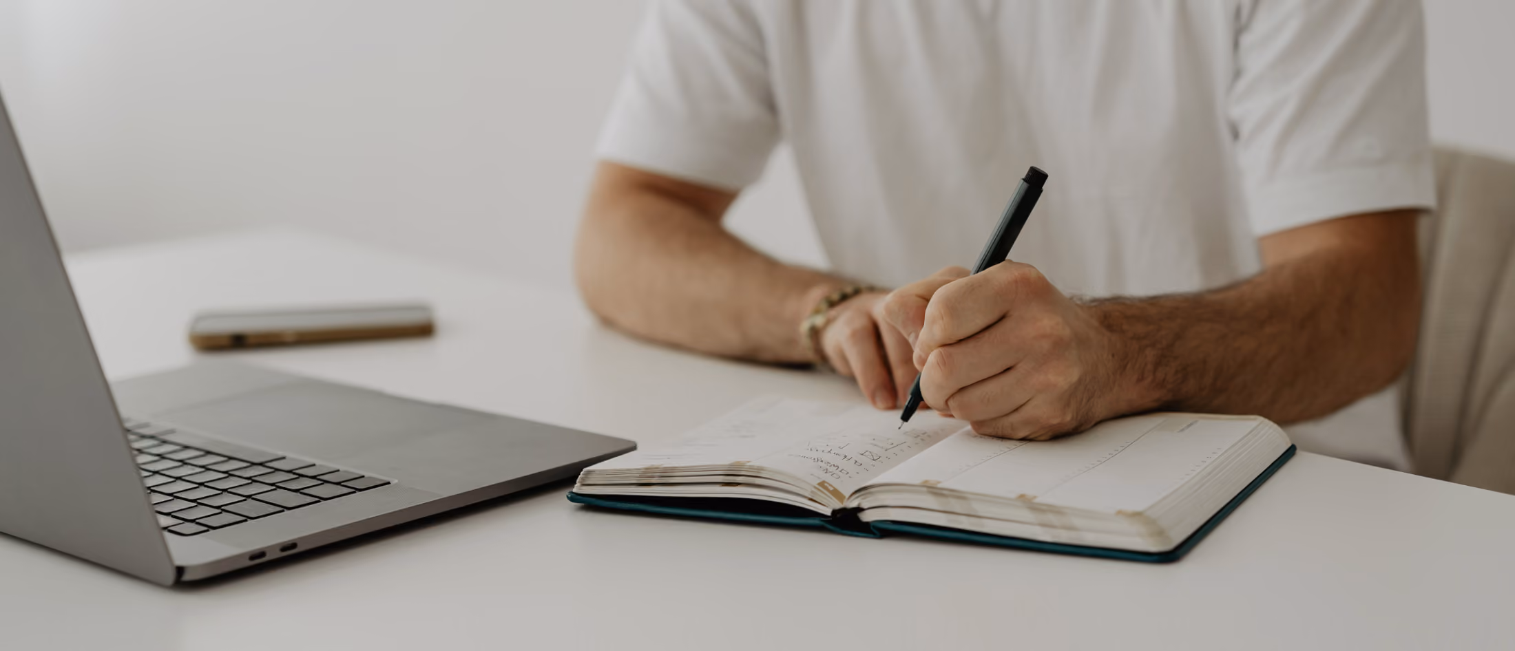 Ein Webdesigner sitzt an einem Schreibtisch mit Laptop und schreibt in ein Notizbuch.
