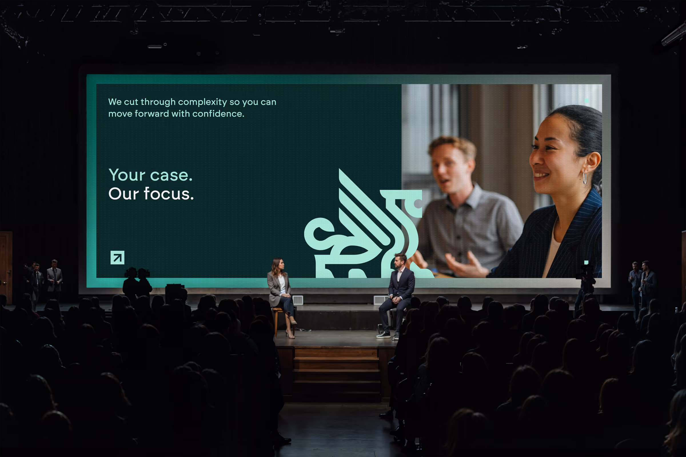 Zwei Moderatoren sitzen auf Stühlen vor einem großen Bildschirm auf einer Bühne vor einem Publikum in einem dunklen Veranstaltungsraum, auf dem Bildschirm steht: 'We cut through complexity so you can move forward with confidence. Your case. Our focus.' mit einem Logo und Bildern von zwei diskutierenden Personen.