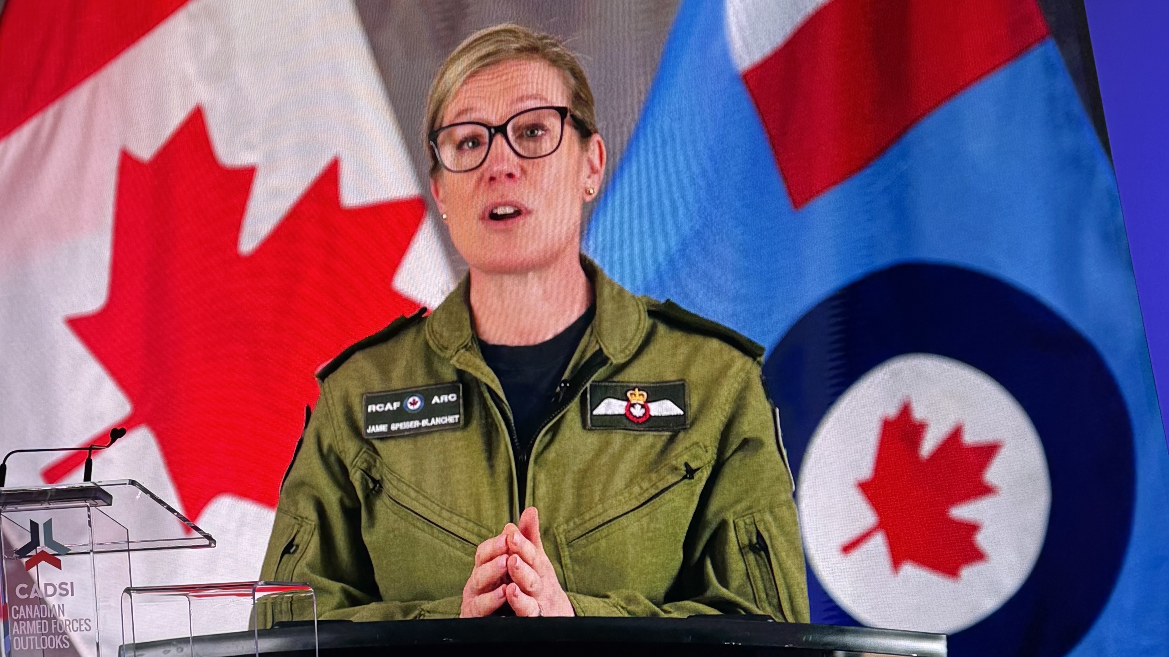 A female general from the Royal Canadian Airforce gives a speech on a podium with Canadian flags behind her.
