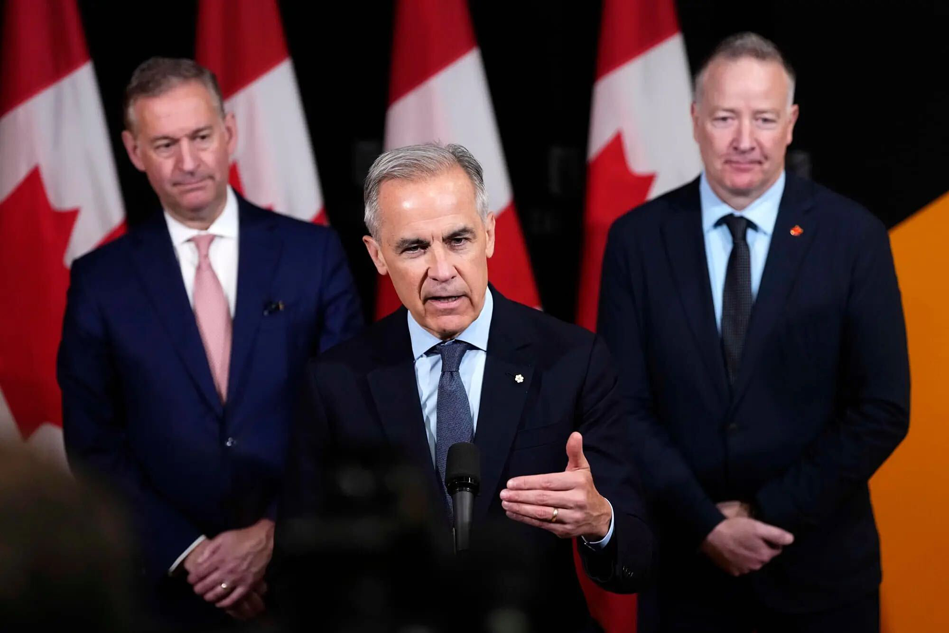 Prime Minister Mark Carney speaks at a podium with two men beside him with Canadian flags behind them