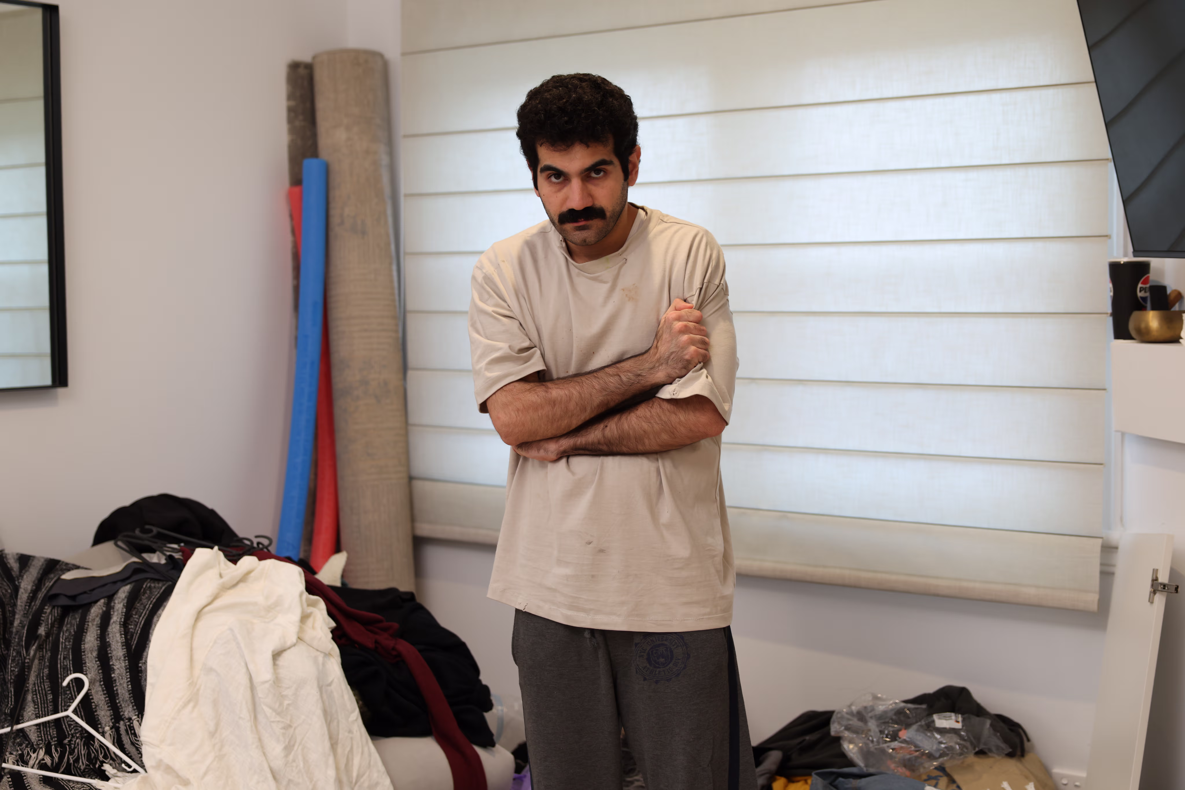A man with a mustache and curly hair stands indoors, wearing a beige shirt and gray pants, with arms crossed and a serious expression.