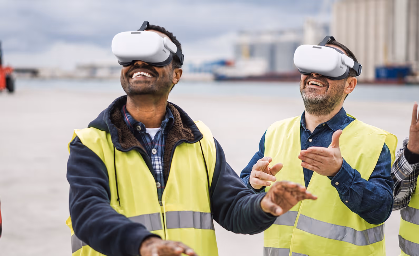 Two warehouse workers in high-visibility vests laughing and gesturing while wearing VR headsets outdoors at an industrial port facility.