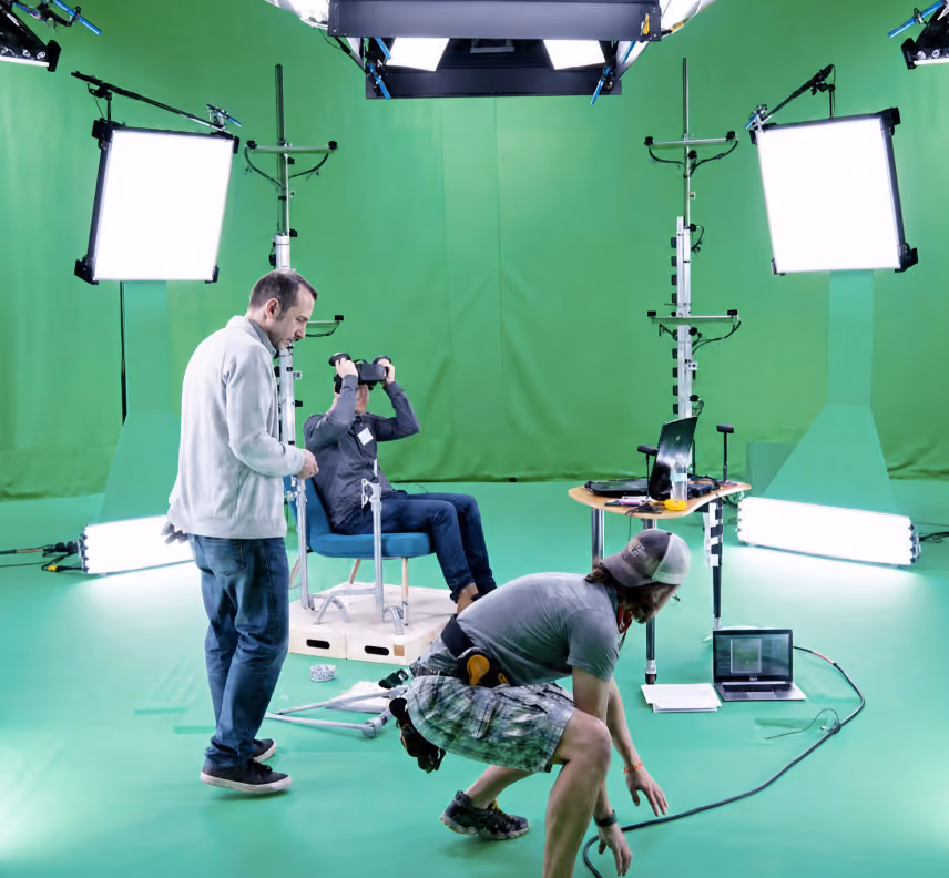 Behind-the-scenes photo of a green screen shoot: crew filming a man wearing a VR headset seated in a rig, with lighting and motion-capture equipment around him.
