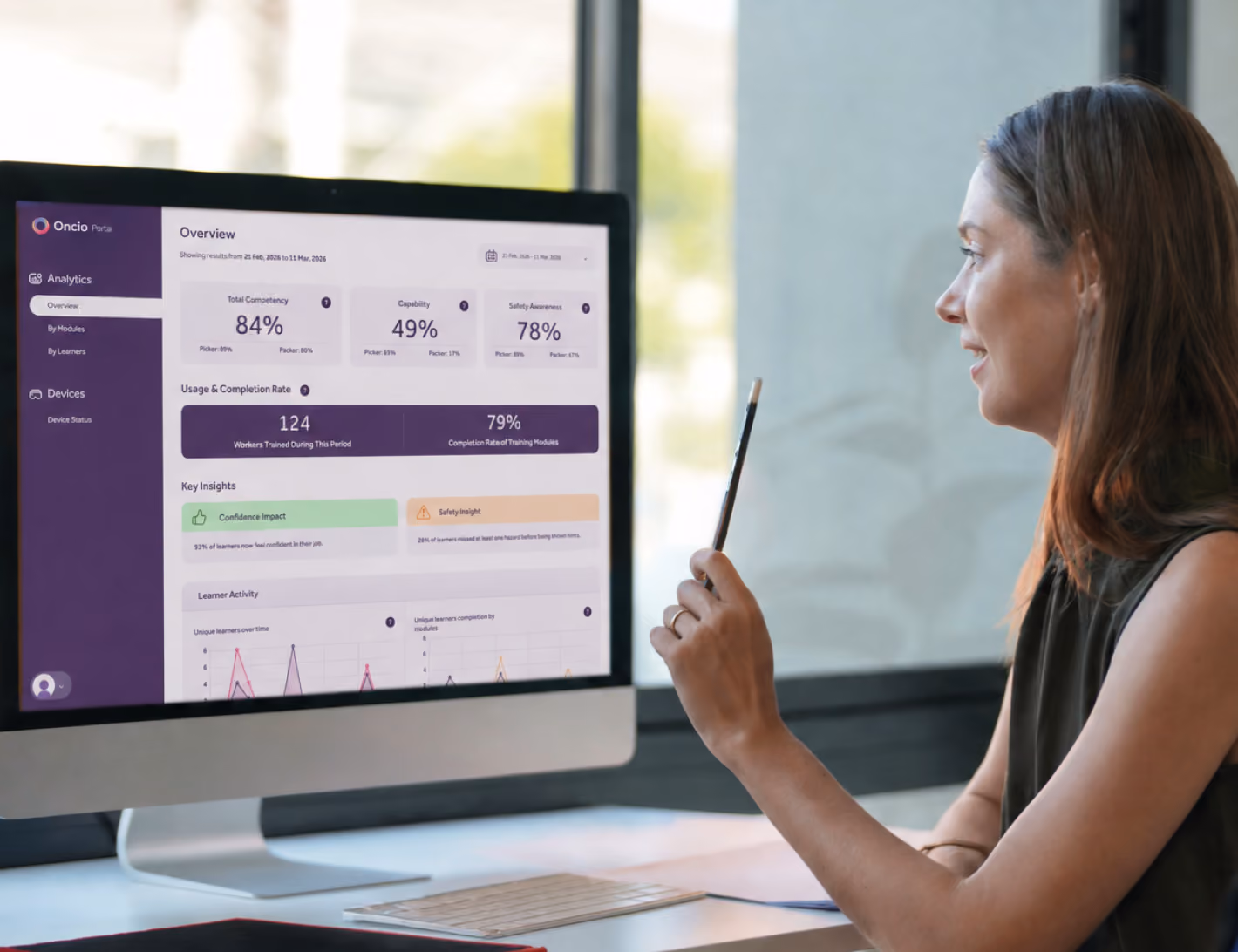 Two women review the Oncio Portal dashboard on a monitor, discussing training completion and competency metrics.