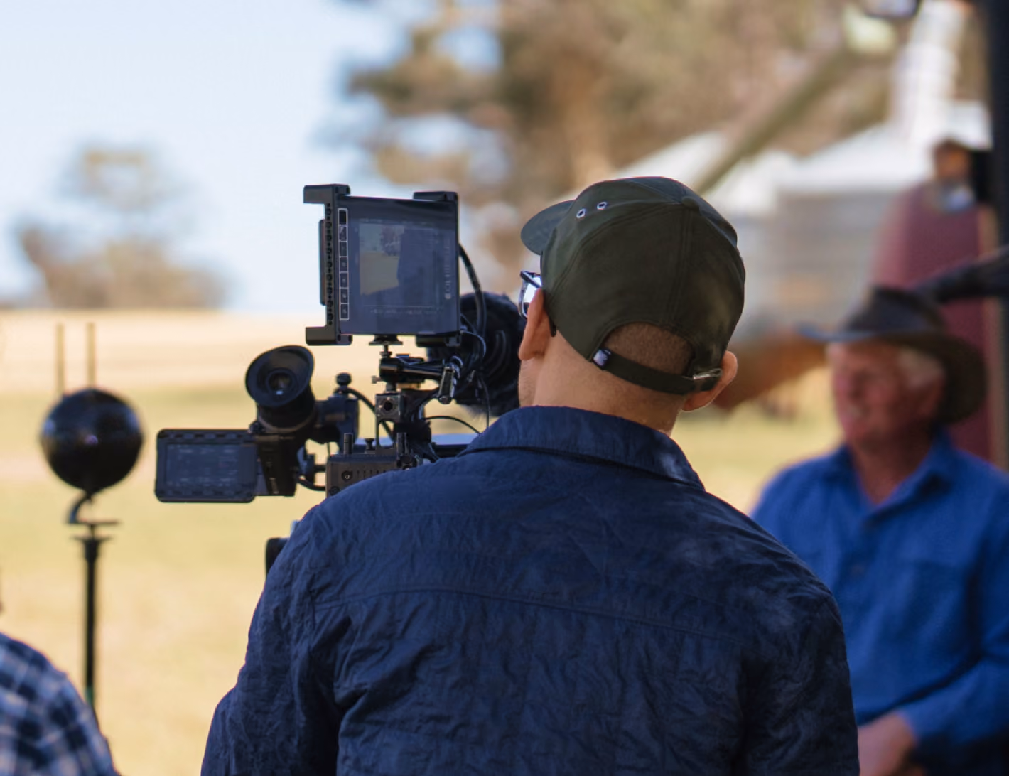 A camera operator films an interview on location with farmers standing beside a red tractor in a paddock.