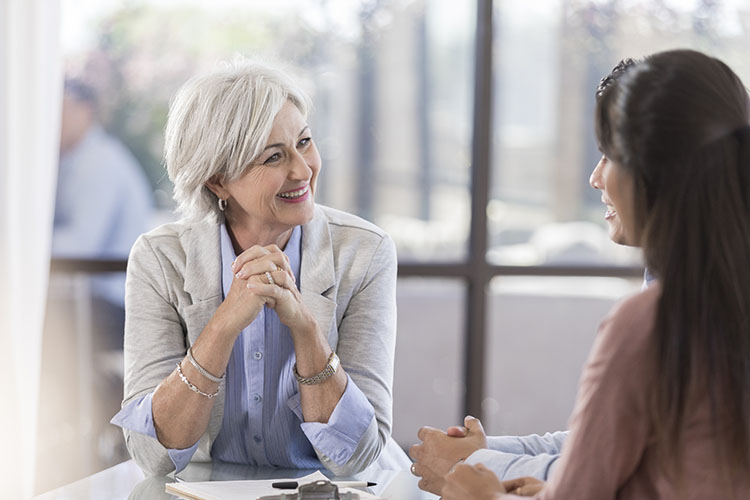 An older business woman with white hair leans forward as she listens intently to another younger woman as she explains something. Social listening is engagingly listening