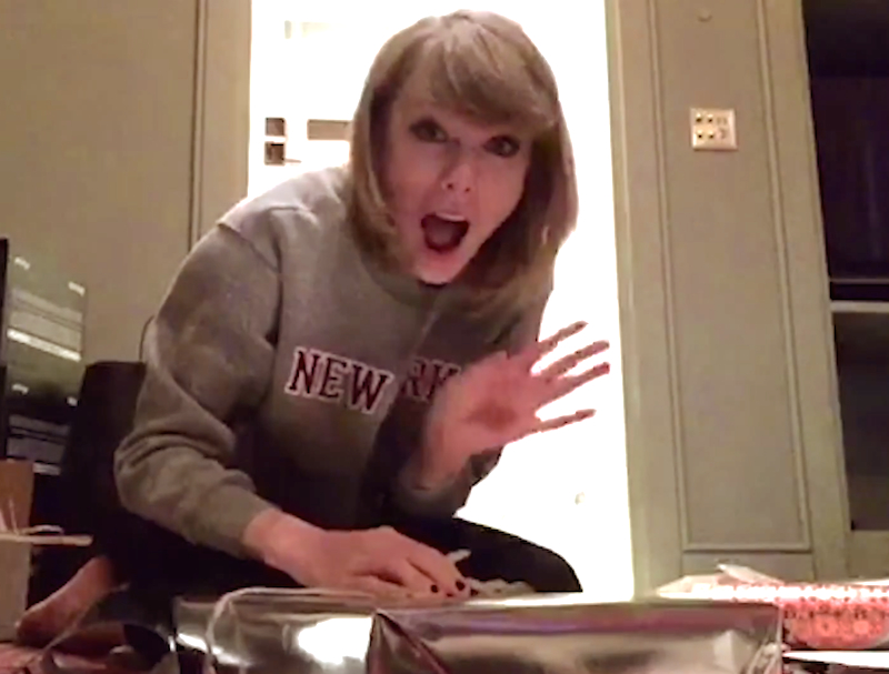Taylor Swift shows people how she did Swiftmas. She is dressed casually in a grey New York sweatshirt. She smiles exaggeratedly at the camera and is waving.