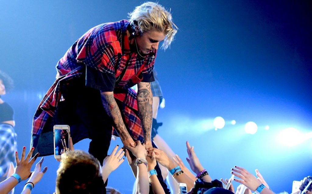 Justine Bieber leans down to hold the hands of a fan. Several more reach out, trying to touch him. Justin is in concert. He is wearing a red and black plaid shirt and tattooed arms. Justin always focused a lot on his fans, knowing that they are what make him successful.