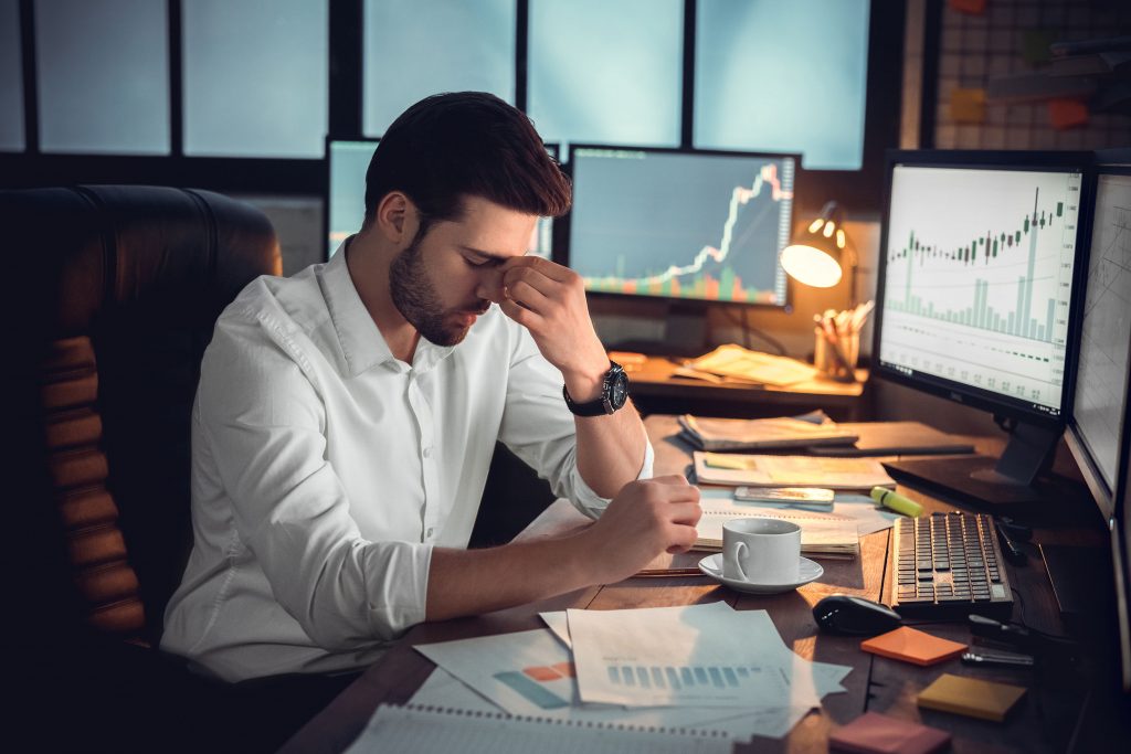 A data scientist at a top consulting firm stressed over the amount of data need to gather an analyze. His desk is cluttered with graphs while his screen has more graphs. He holds his nose as he takes a short mental destressing moment.