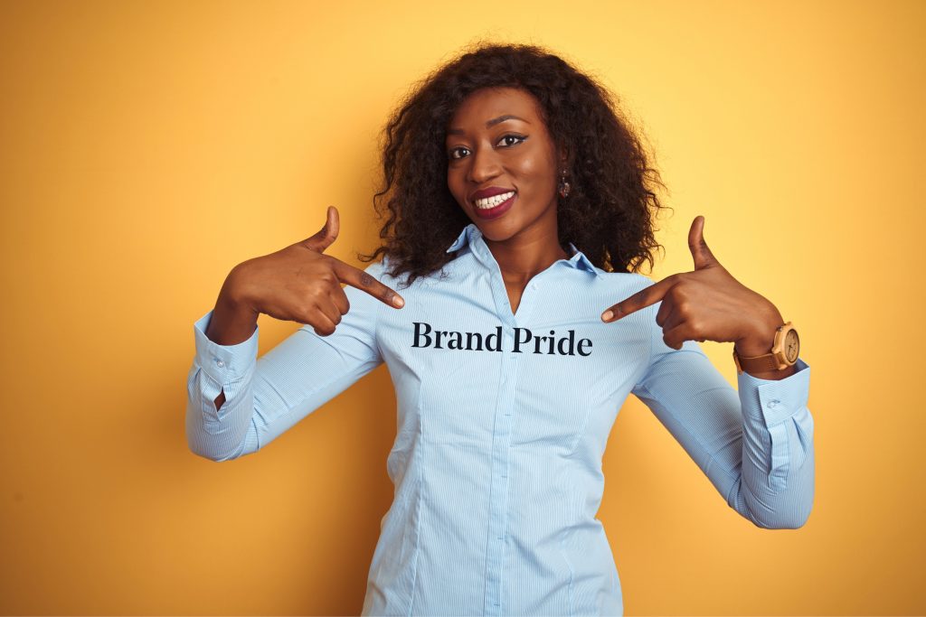 A black woman in a light blue collared shirt has both hand level with her shoulders and points at her chest. She is smiling and her head is slightly tilted to the left. Across her chest is written Brand Pride.