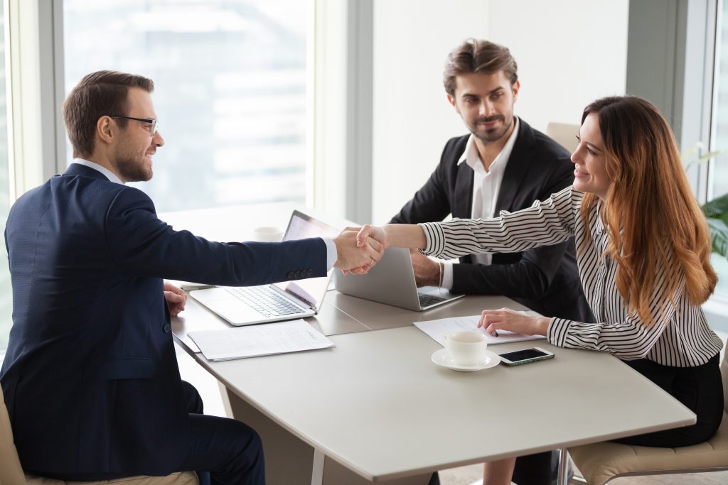 Three people in business attire sit at a table. Two are white men and the third is a woman. The woman shakes the hand of the man sitting diagonally across the table from her. The second man sits directly across the table from the man she is shaking hands with. When companies implement strategies that focus on customers, customer are happy., just like this smiling woman.