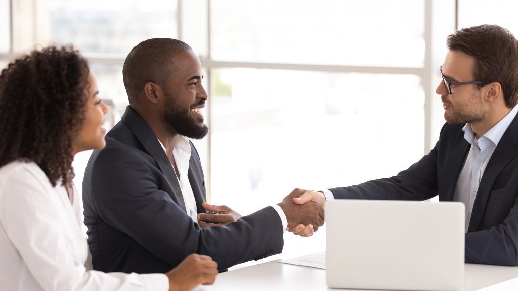 Three people in business attire sit at a table. Two are men and one is a woman. The woman appears to be Latino, the man directly to her left is black, and the man sitting at across the table is white. The black man and white man shake hands. The black man and woman smile happily at the white man.