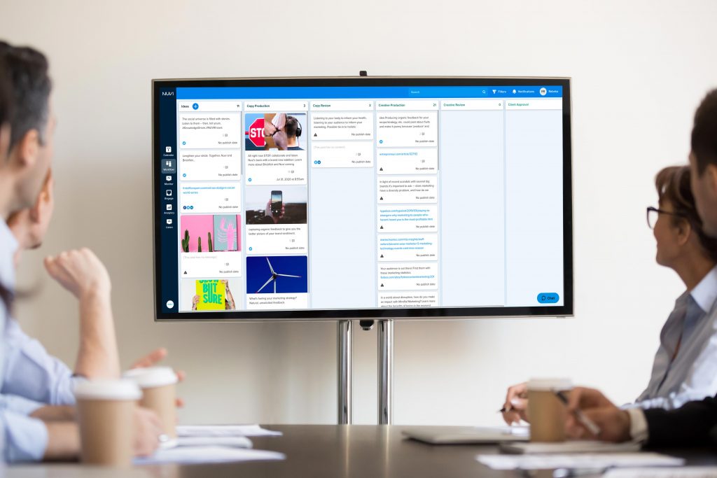 A group of people sit around a conference table. The look at a screen that has Nuvi Plan on it. There are seven KANBAN-workflow columns that content can be approved and moved through to publishing. This is especially good for user-generated content.
