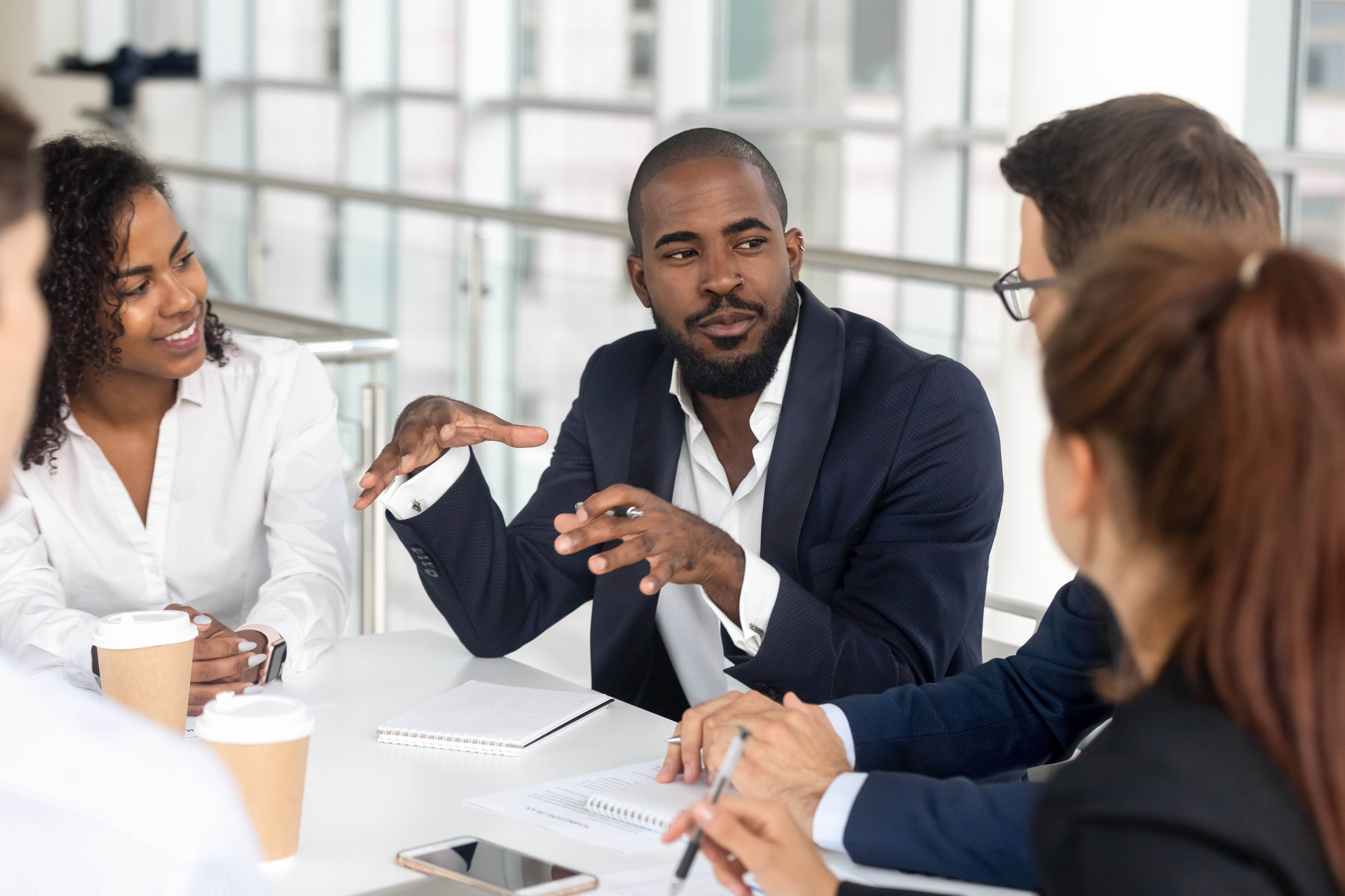 An older man, late 30's, is training a group of interns or new employees. His hands are held out in front of him while he explains something. The younger people watch intently.