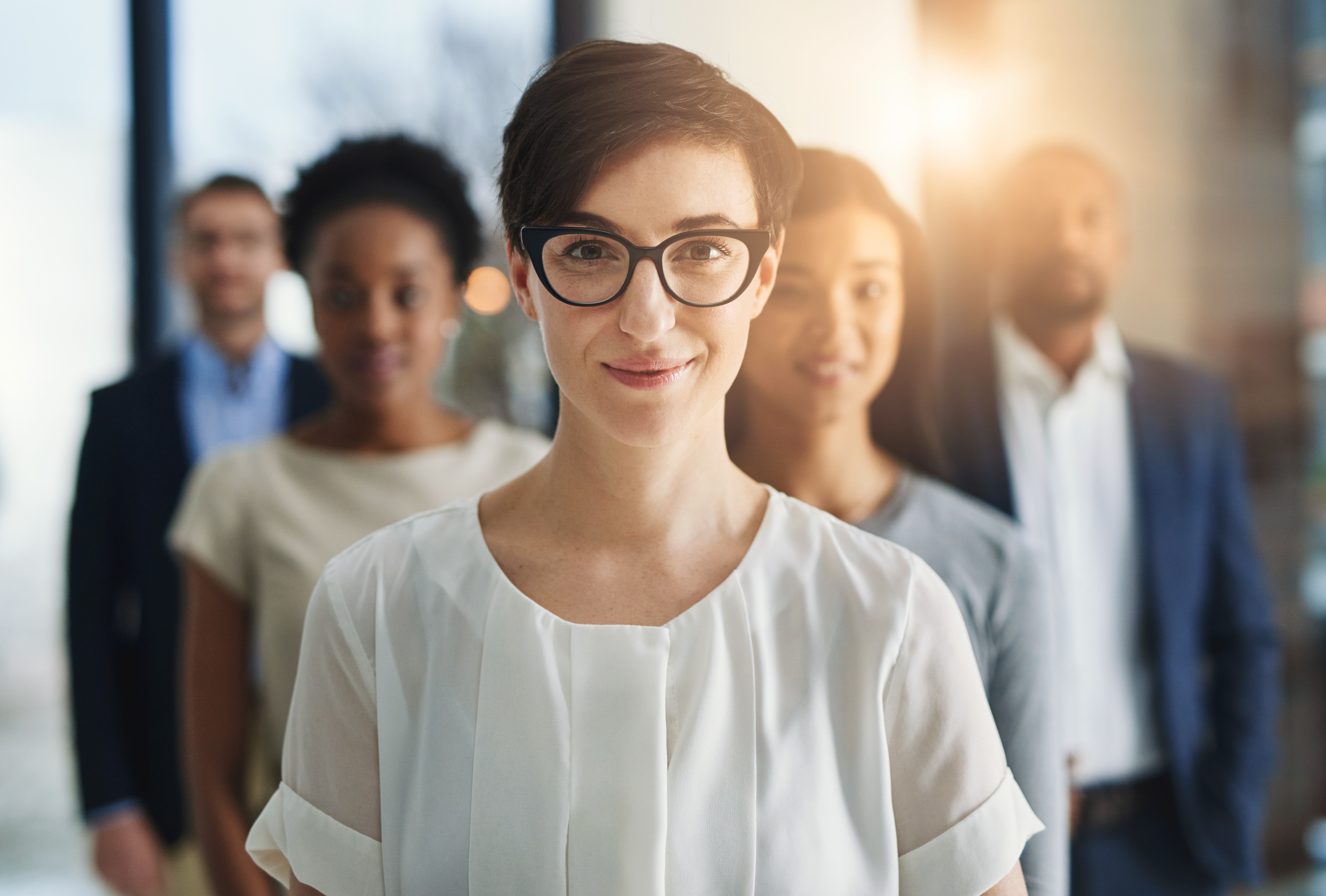 Five employees stand in a triangle, the point facing the camera. They all stand confidently. The woman in front is smiling confidently, two other women stand behind her, and two men in casual work suits stands behind them.