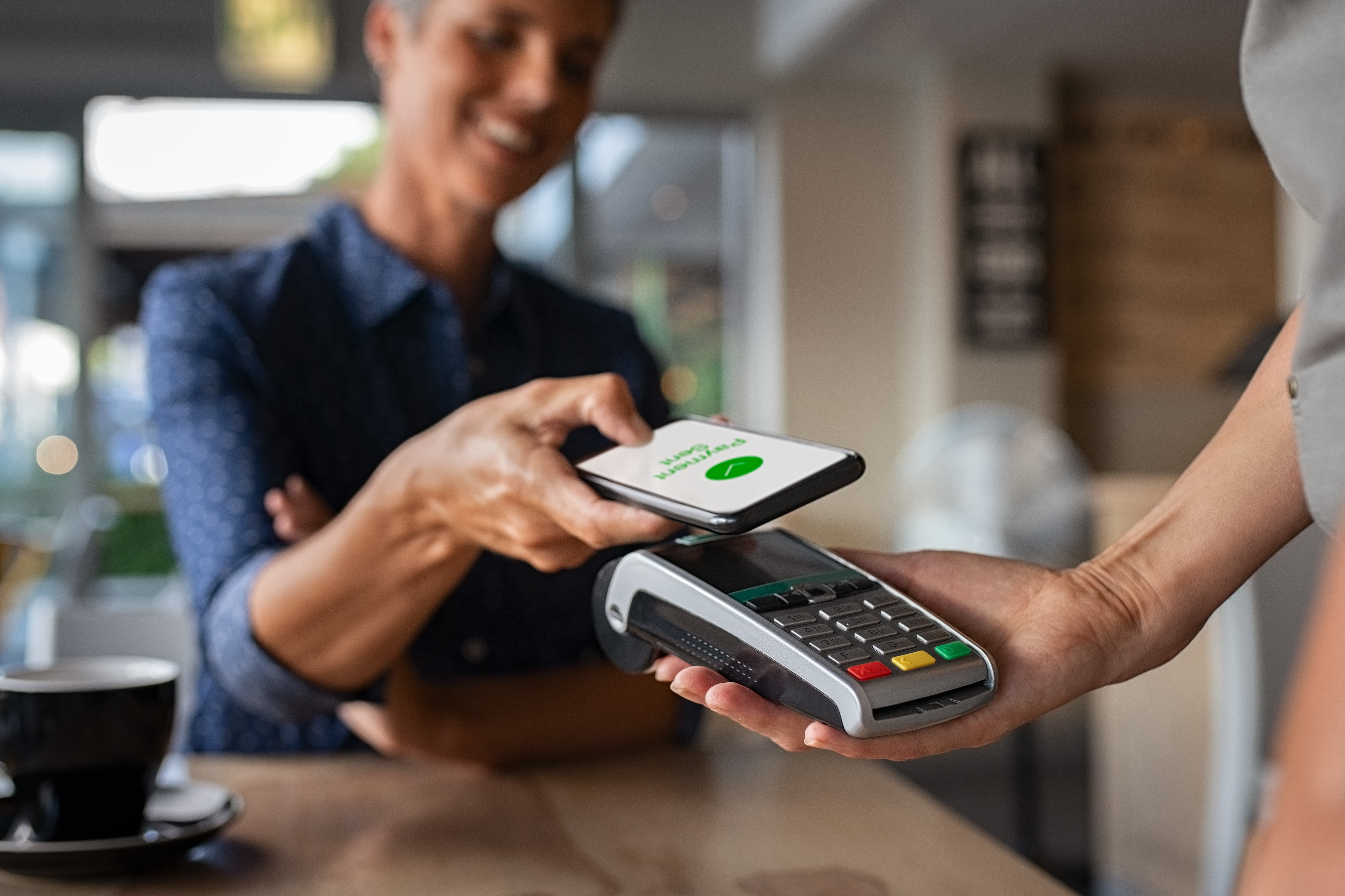 A customer, who is blurred, holds his or her phone over the payment device that the employee is holding up. The phone has a green circle with a check in it over the words Payment Sent. Getting a customer to purchase, then cross sell or up sell them is a challenge with how easily customers can be informed now.