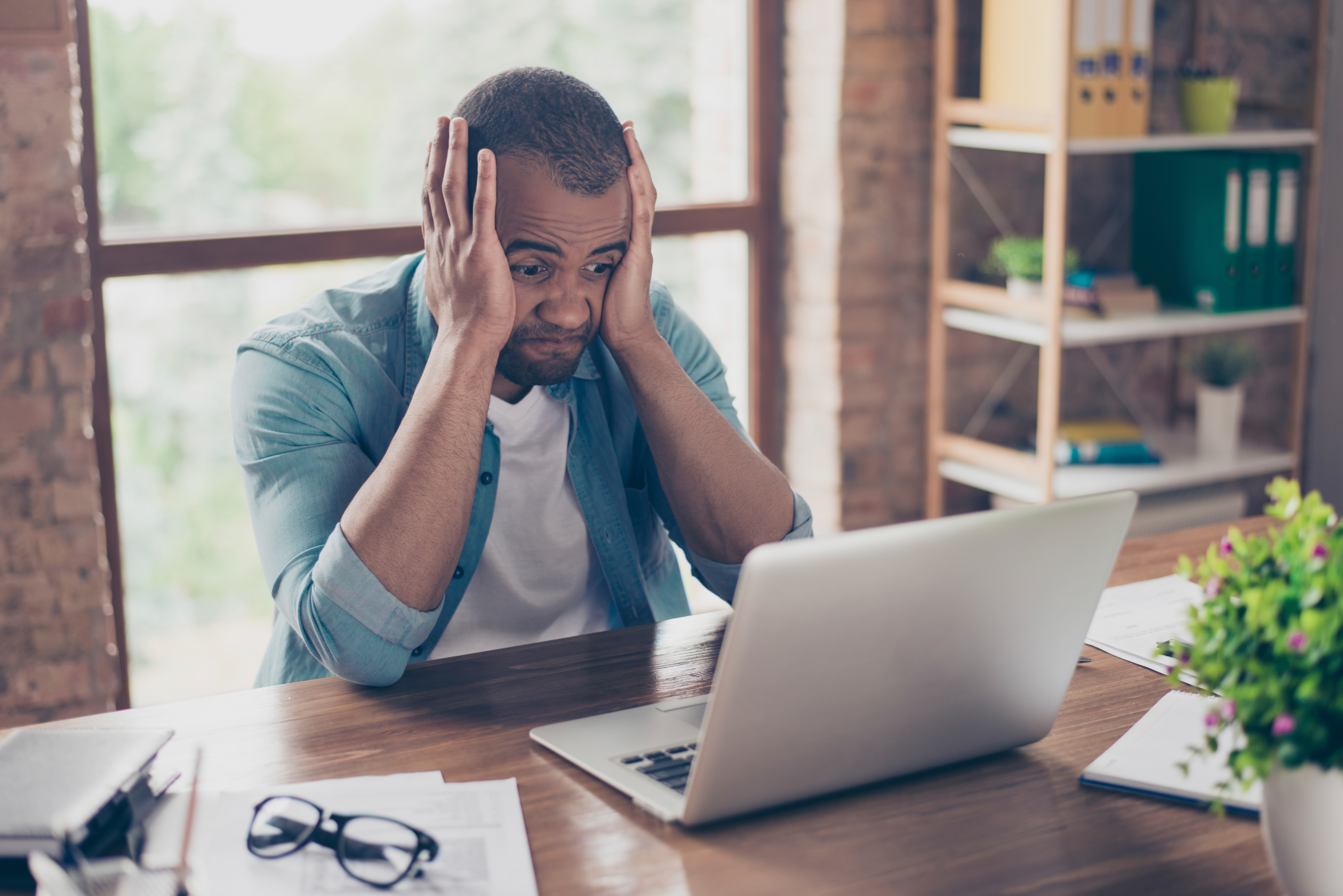 A man working in an bricked office holds his head between hands as he looks at his computer in frustration. His elbow are in his desk as he leans forward. Getting stuck in a routine can lead to the biggest mistakes.