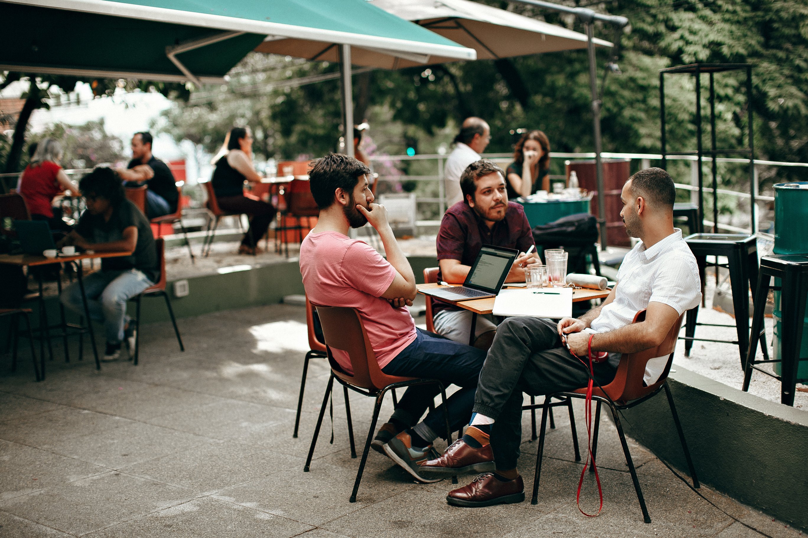 Many people sit out round tables at an outdoor restaurant. The tables are covered with square awnings. The images is focused on three men at a table together. The are having a friendly chat and enjoying time together.