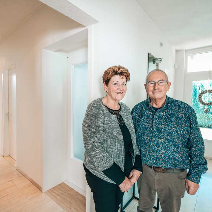 Een vrolijk ouder echtpaar, een vrouw met kort rood haar en een man met een bril, glimlacht in de lichte gang voor hun huislift