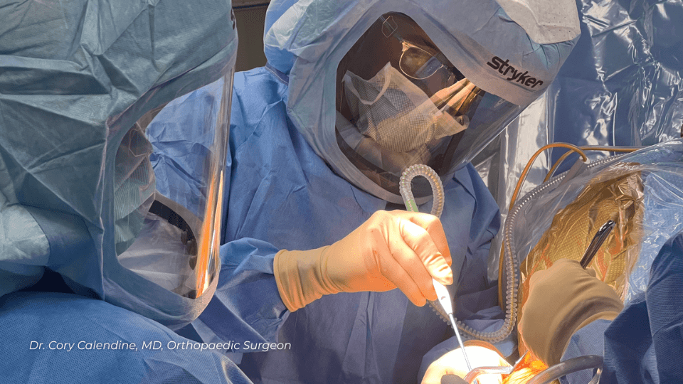 Dr. Cory Calendine performing hip replacement surgery in sterile operating room at Williamson Medical Center Franklin Tennessee, wearing surgical mask and protective equipment while conducting orthopedic procedure