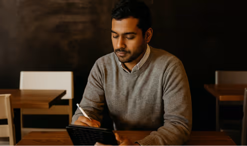 Man in a gray sweater writing on a notepad while sitting at a wooden table in a dimly lit room.